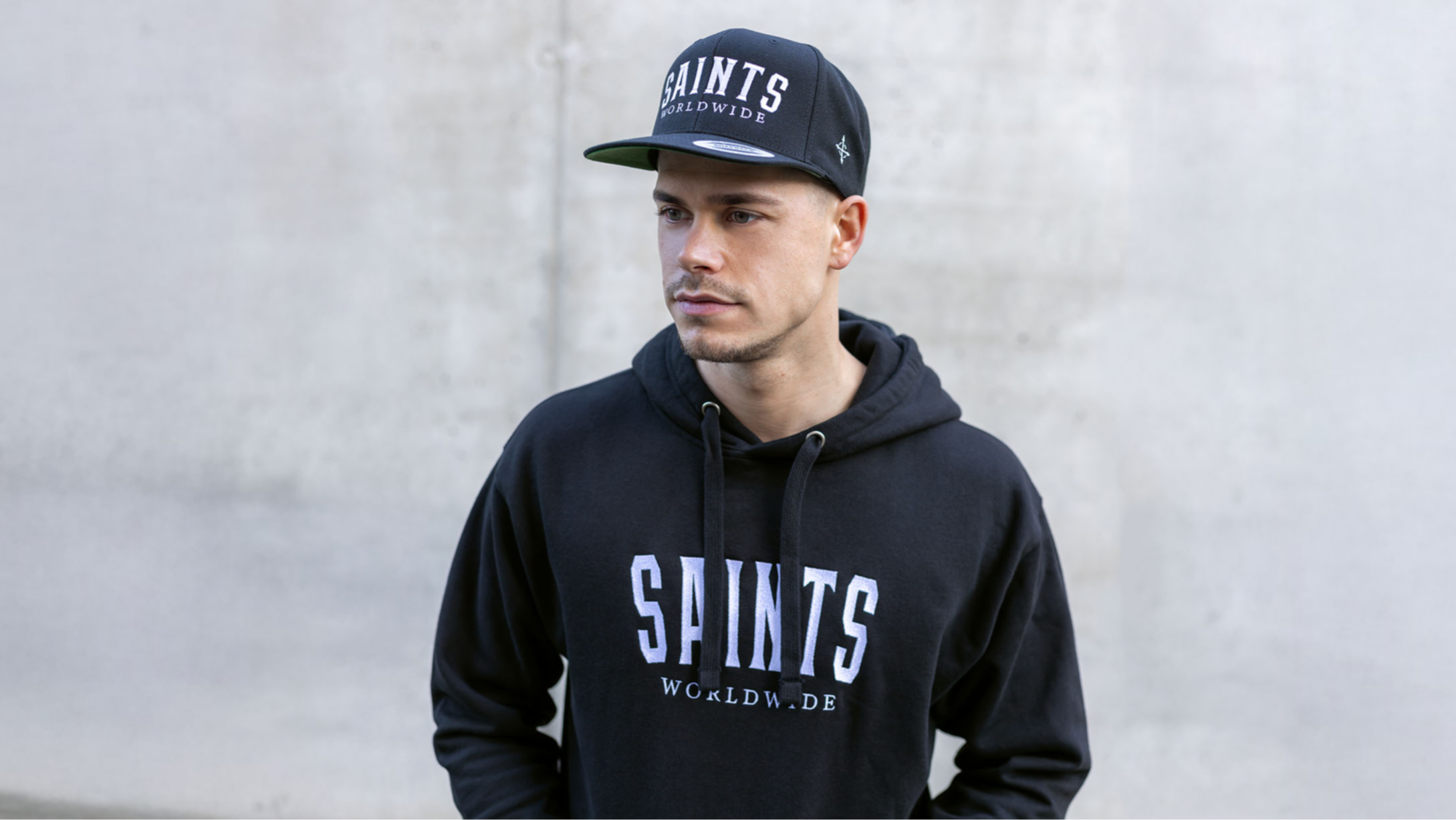 Model wearing Saints Worldwide Christian hoodie and snapback against light concrete background